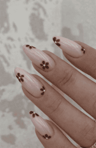 Almond-shaped nude nails with subtle vertical texture and small chocolate brown daisy flower accents, glossy finish with soft minimalist floral nail art