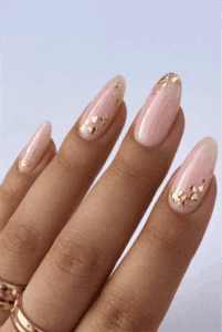Medium almond nails with blush pink nude base and scattered gold metallic foil accents, glossy finish with soft romantic luxury nail design