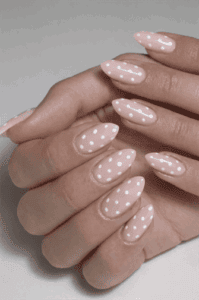 Almond-shaped nails with soft blush pink base and small white polka dot pattern, glossy finish with playful minimalist aesthetic and clean modern nail design