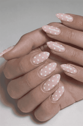 Almond-shaped nails with soft blush pink base and small white polka dot pattern, glossy finish with playful minimalist aesthetic and clean modern nail design