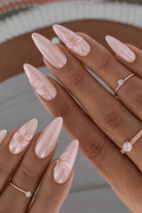 Long almond-shaped nails with soft pink pearlescent chrome finish and delicate 3D flower accents, glossy iridescent shine with elegant bridal-inspired floral nail design