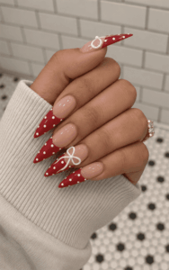 Red polka dot French tip stiletto nails with white 3D bow accents, glossy nude base and festive coquette-inspired nail design
