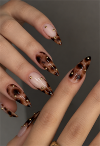 Long stiletto nails with tortoise shell tips, sheer nude accents, gold studs, and a glossy finish