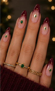 Long almond nails with burgundy French tips, emerald green rhinestones and delicate gold starburst accents.